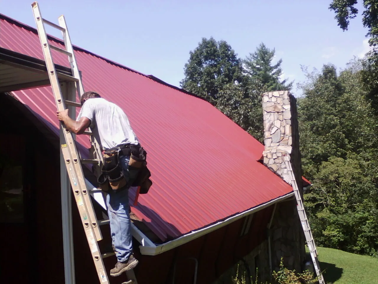 Roofer on a ladder beside a red metal roof and stone chimney — Soffit & Fascia Repair by Roofing Brothers in Buhler